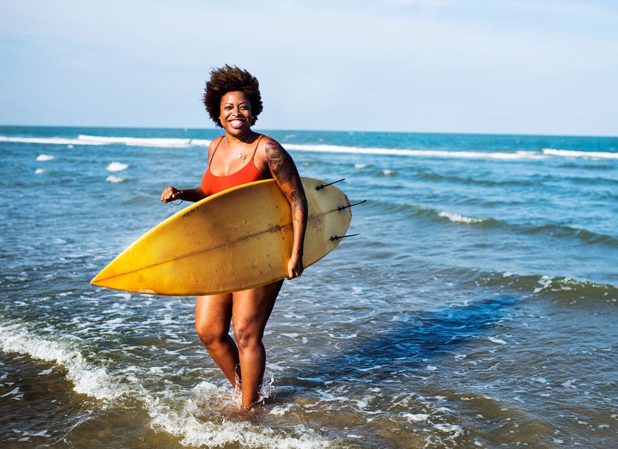 Middle aged woman on the beach with a surfboard smiling at the camera